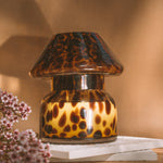 Mushroom candle lamp with light and dark brown spots on tan coloured glass. Leopard candle lamp is filled with 100% soy wax sitting on white tiles next to dried flowers.