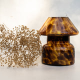 Mushroom candle lamp with light and dark brown spots on tan coloured glass. Leopard candle lamp is filled with 100% soy wax next to dried flowers.