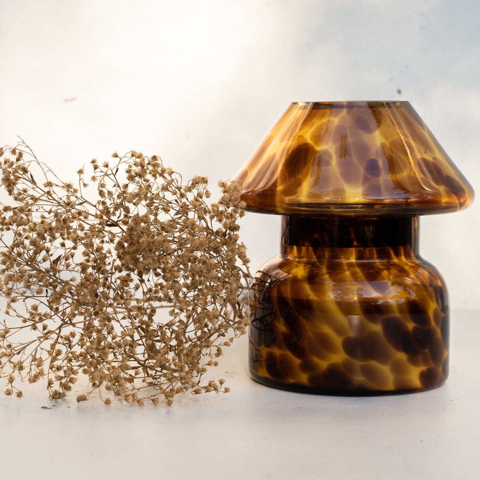 Mushroom candle lamp with light and dark brown spots on tan coloured glass. Leopard candle lamp is filled with 100% soy wax next to dried flowers.