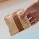 Hand holding a beige product packaging with a brown label against a neutral background. Herbal bath soak.