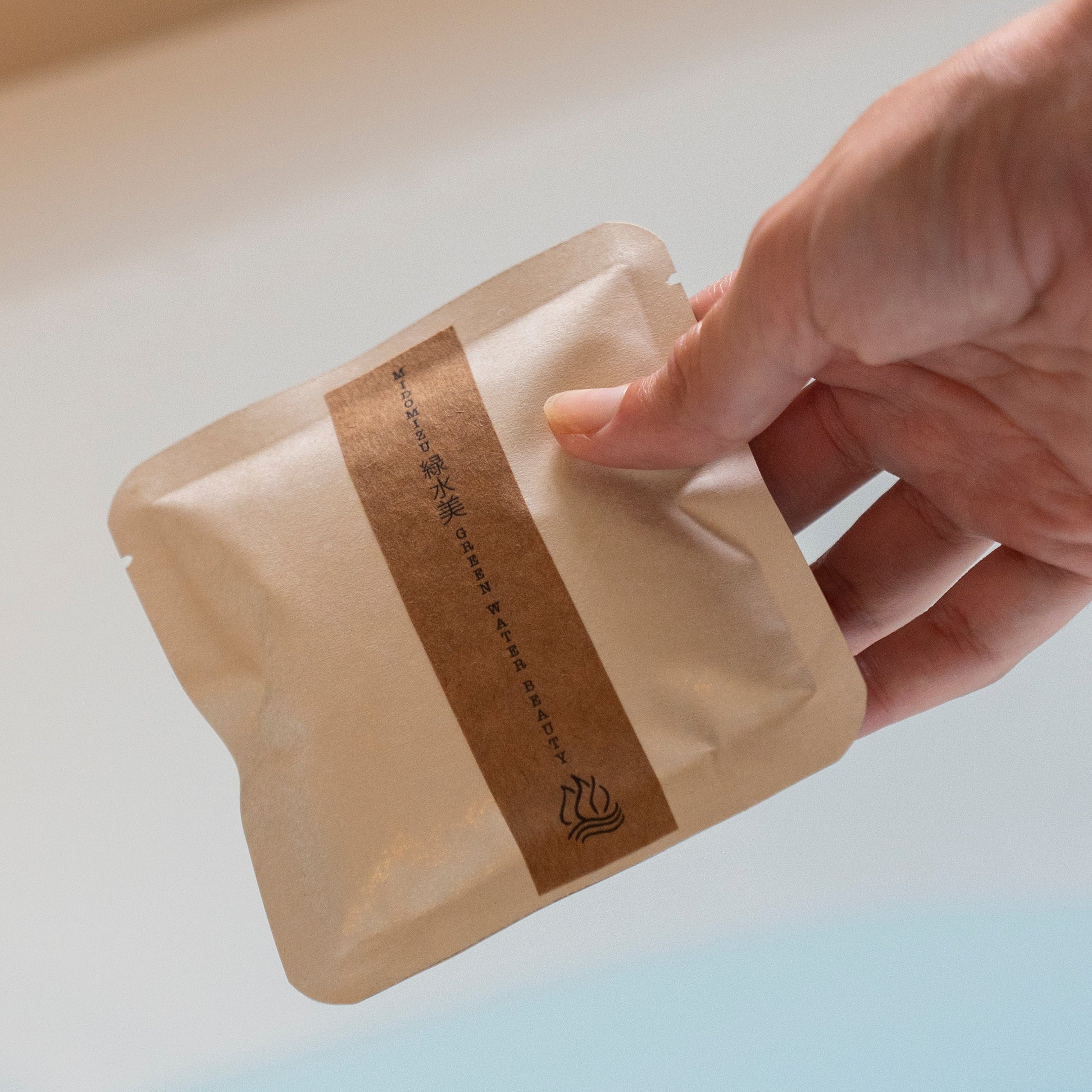 Hand holding a beige product packaging with a brown label against a neutral background. Herbal bath soak.