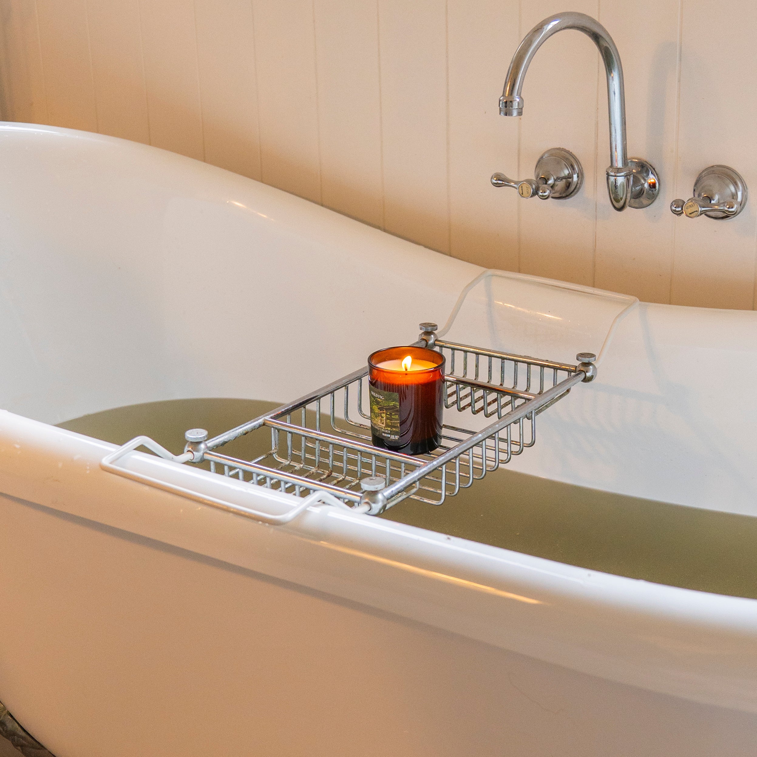 White freestanding bathtub with a metal tray holding a candle, against a beige tiled wall.