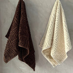 Two textured hand towels in brown, beige, and green hanging on a gray wall.