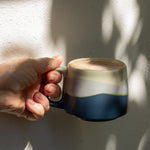 Hand holding a ceramic mug with a layered design against a textured wall.