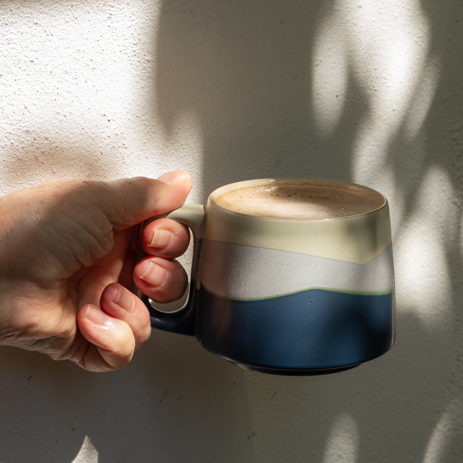 Hand holding a ceramic mug with a layered design against a textured wall.