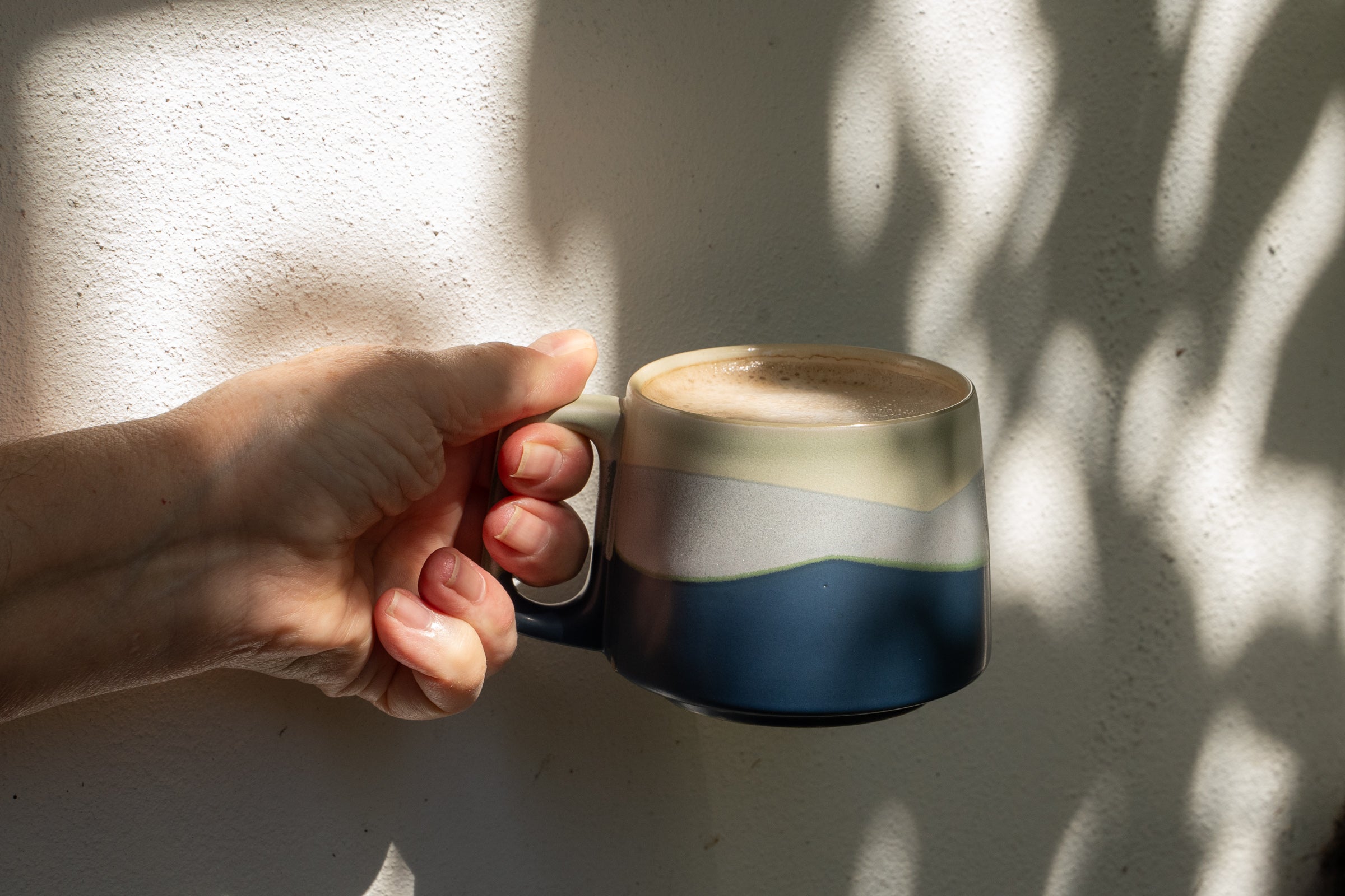 Hand holding a ceramic mug with a layered design against a textured wall.