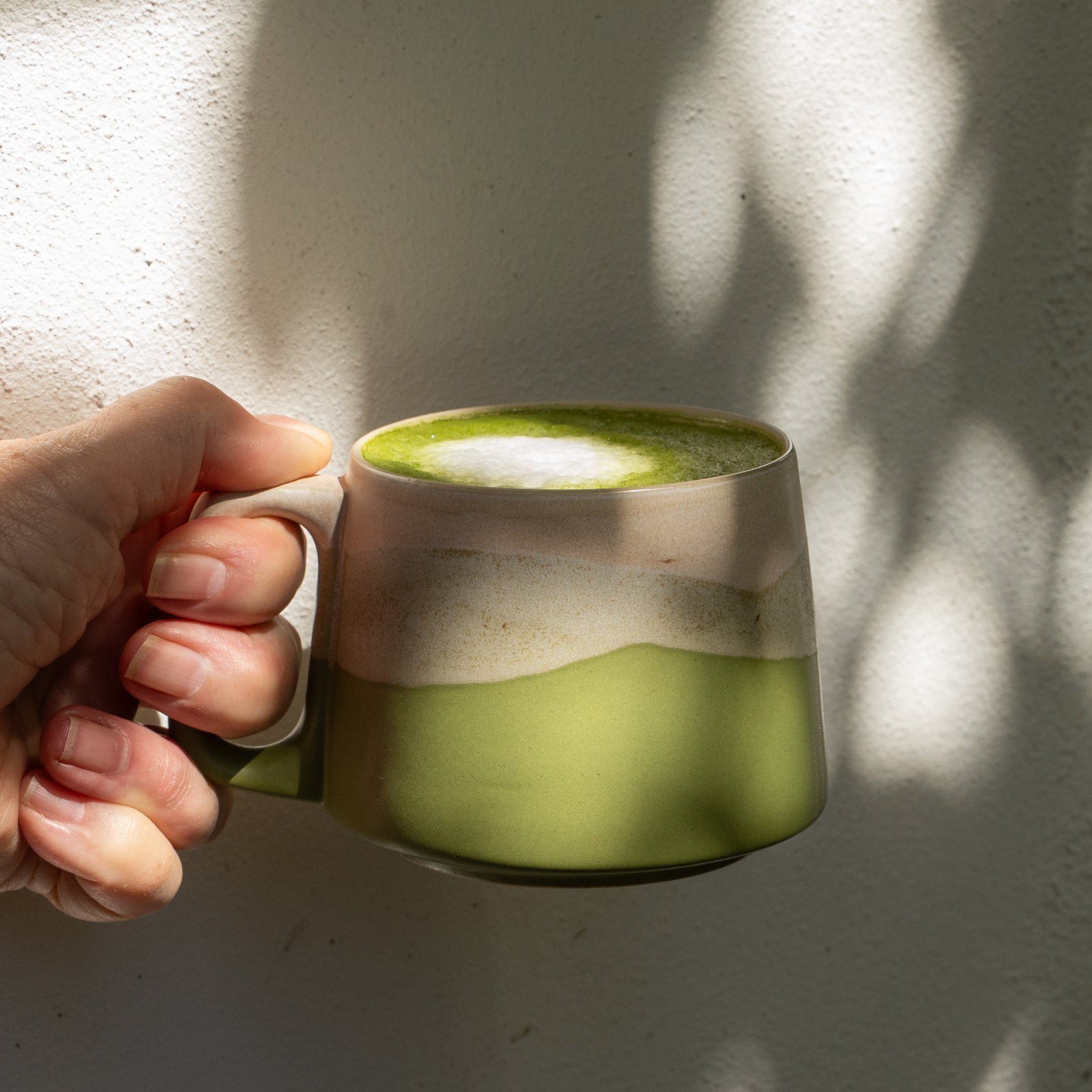 Hand holding a green mug with a creamy beverage against a textured wall.