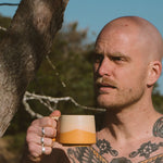 Man with tattoos holding a mug outdoors near a tree
