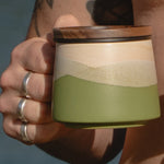 Hand holding a ceramic mug with a layered design, featuring green, beige, and brown colors.