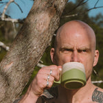Man with tattoos drinking from a green mug outdoors near a tree