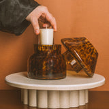 Person lighting a candle in a tortoiseshell-patterned glass holder on a small round table against an orange wall.