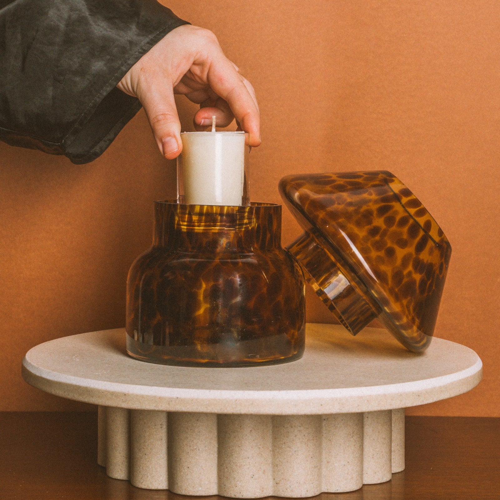 Person lighting a candle in a tortoiseshell-patterned glass holder on a small round table against an orange wall.