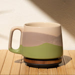 Ceramic mug with a landscape design on a wooden surface with a shadow of a plant.