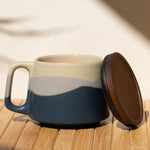 Ceramic mug with a lid on a wooden surface with a leaf shadow on the wall.