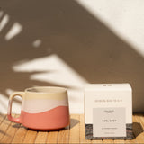 Pink and beige ceramic mug with a box of Earl Grey tea on a wooden surface with a white wall background.