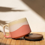 Ceramic mug with a pink and beige design on a wooden surface with a shadow cast by a plant.