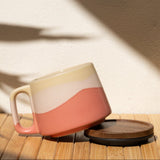 Ceramic mug with a pink and beige design on a wooden surface with a shadow cast by a plant.