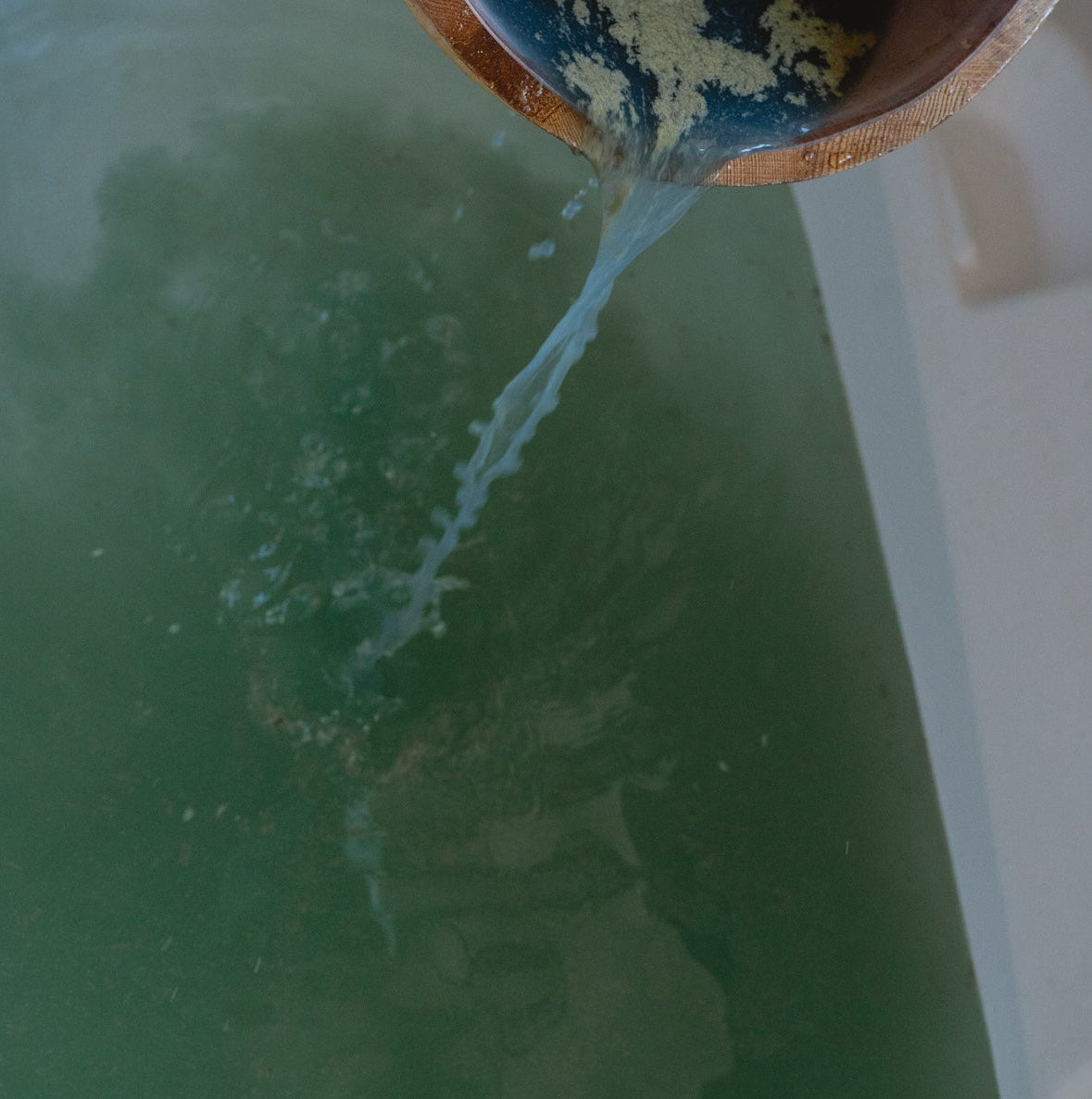 Person pouring water from a wooden bucket into a bathtub filled with herbal bath product