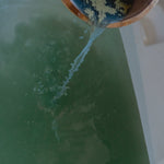 Person pouring water from a wooden bucket into a bathtub filled with herbal bath product
