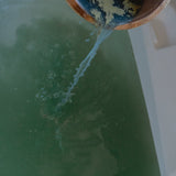 Person pouring water from a wooden bucket into a bathtub filled with herbal bath product
