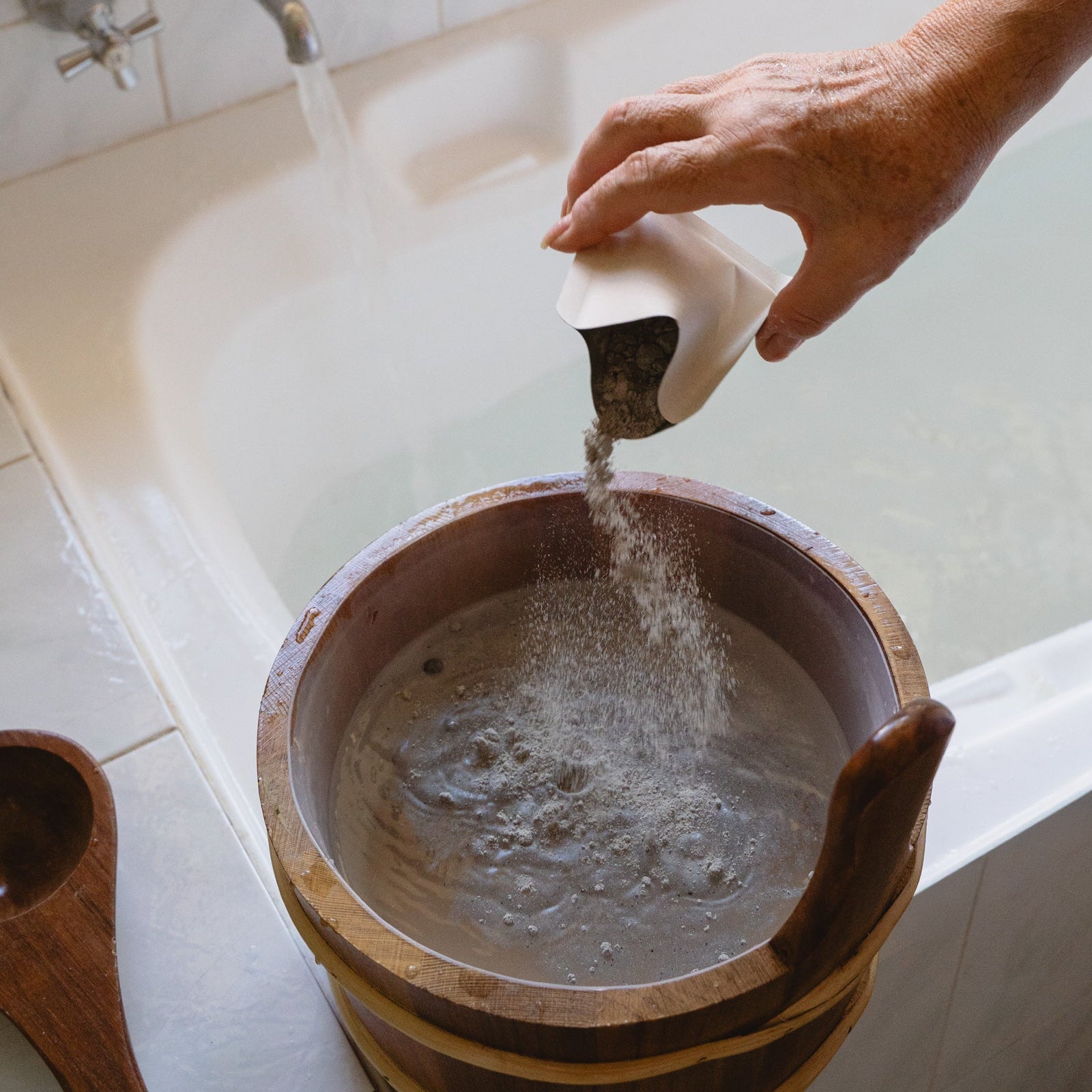 Person pouring herbal bath soak into a wooden bucket in a bathroom.