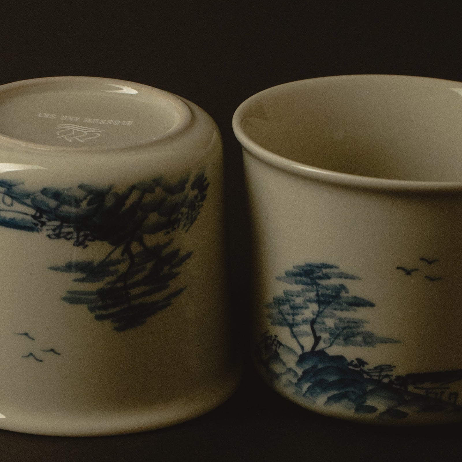 Two ceramic cups with blue tree and bird designs on a dark background