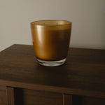 An amber glass candle jar with a clear glass base, sitting on a dark wooden surface against a plain light gray wall.