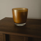 An amber glass candle jar with a clear glass base, sitting on a dark wooden surface against a plain light gray wall.