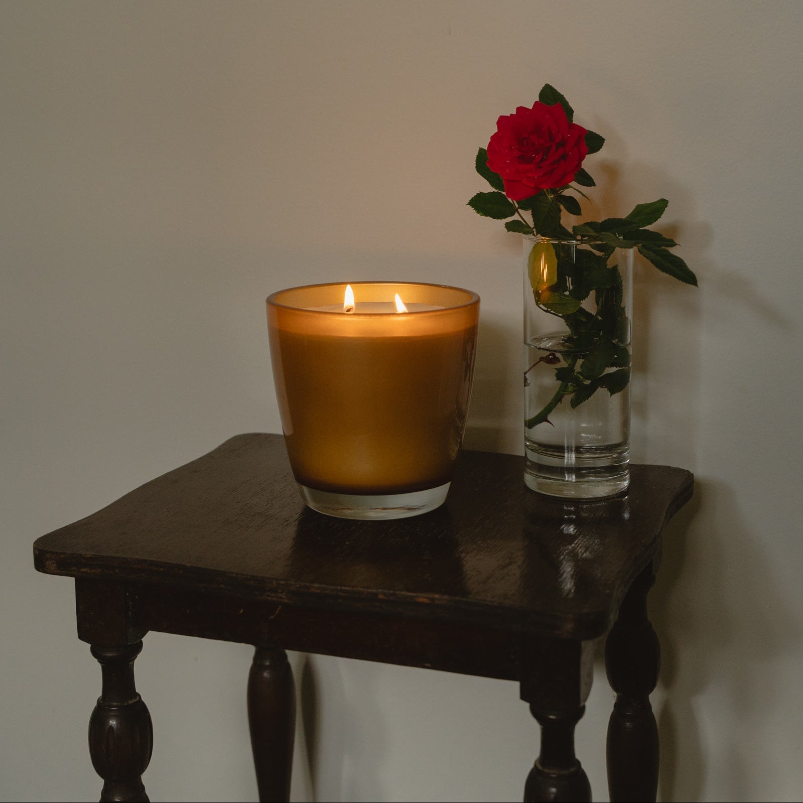 A lit amber glass candle sits on a dark wooden table next to a clear glass vase holding a single red rose, against a plain light wall.