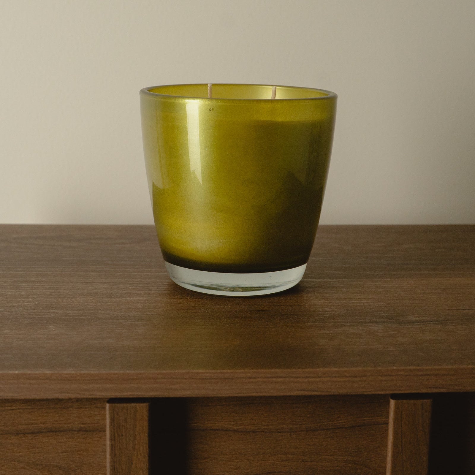 An olive green glass candle holder with a clear glass base, sitting on a dark wood table against a plain beige wall.