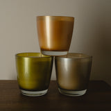 Three frosted glass candle holders in amber, olive green, and champagne tones, stacked in a pyramid on a dark wooden surface against a neutral beige background.