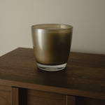 A frosted glass candle holder with a warm bronze-brown tint, sitting on a dark wooden table against a neutral beige wall.