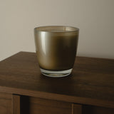 A frosted glass candle holder with a warm bronze-brown tint, sitting on a dark wooden table against a neutral beige wall.