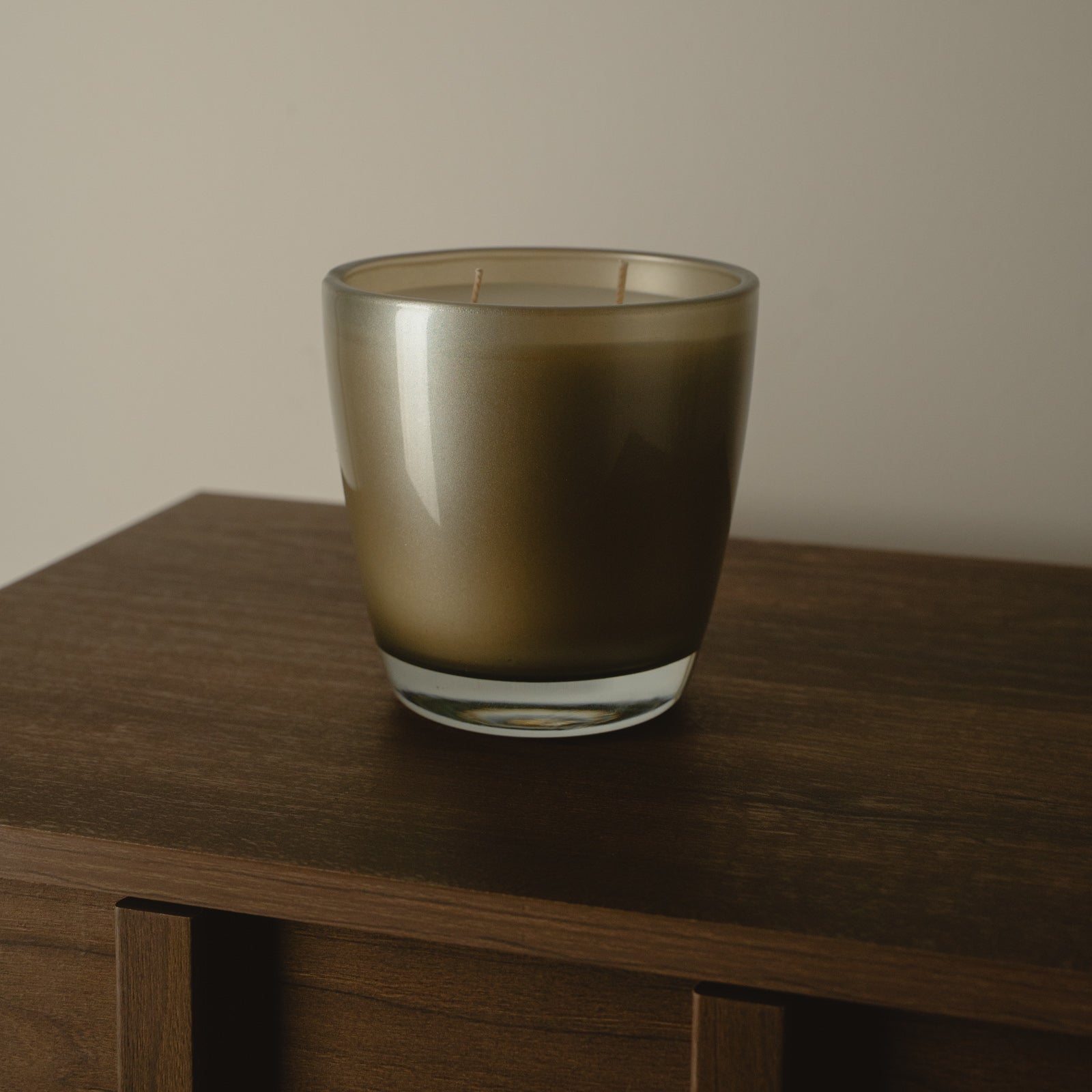 A frosted glass candle holder with a warm bronze-brown tint, sitting on a dark wooden table against a neutral beige wall.