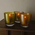 Three amber glass candle holders with a warm glow from lit candles inside, arranged on a dark wooden surface against a neutral beige wall.