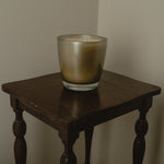 A two-wick candle in a frosted glass holder with a metallic gold-to-silver gradient, displayed on a dark wooden side table against a plain beige wall.
