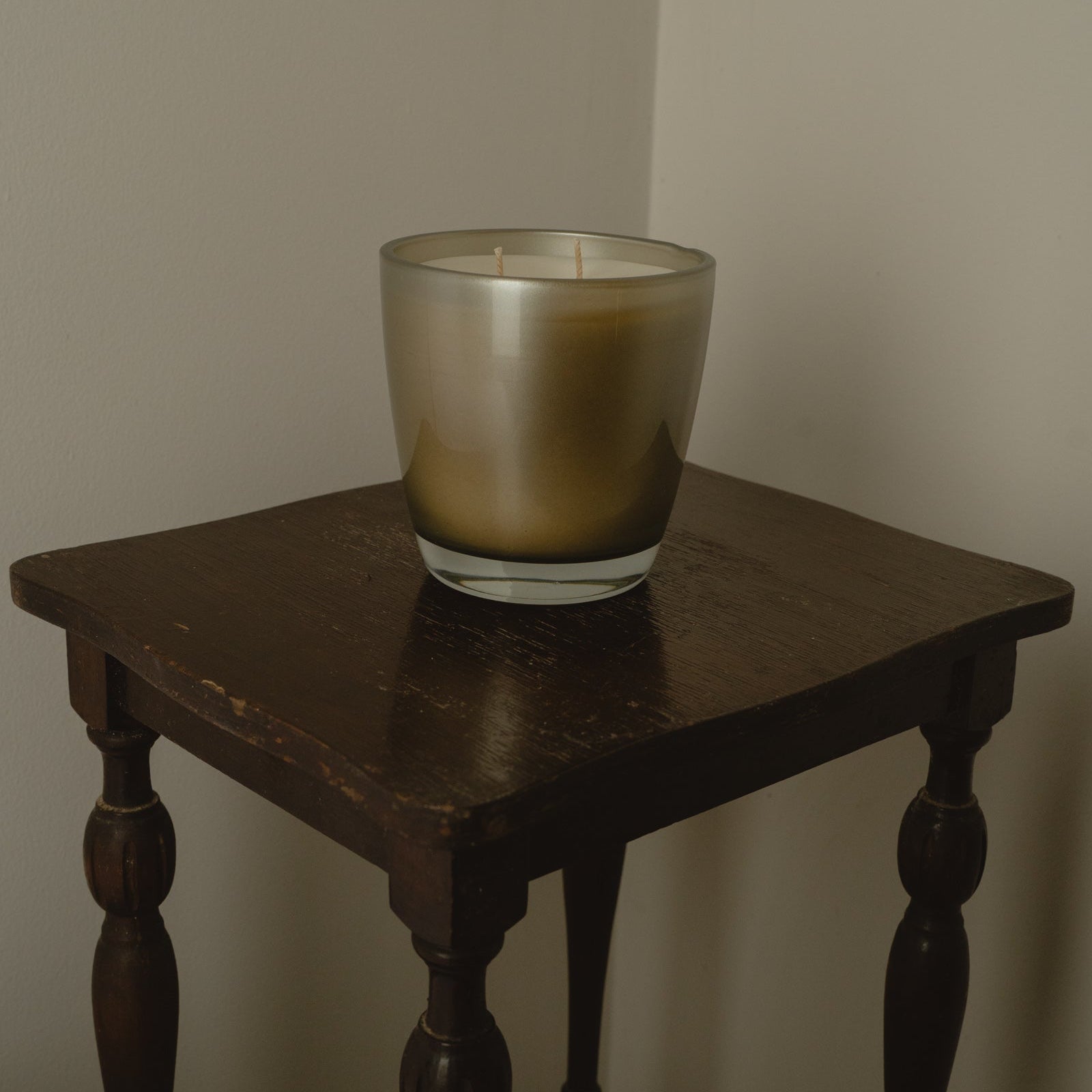 A two-wick candle in a frosted glass holder with a metallic gold-to-silver gradient, displayed on a dark wooden side table against a plain beige wall.