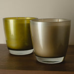 Two glass candle holders on a wooden surface against a neutral beige wall. The left holder is olive green, and the right holder is a smoky taupe-brown gradient.