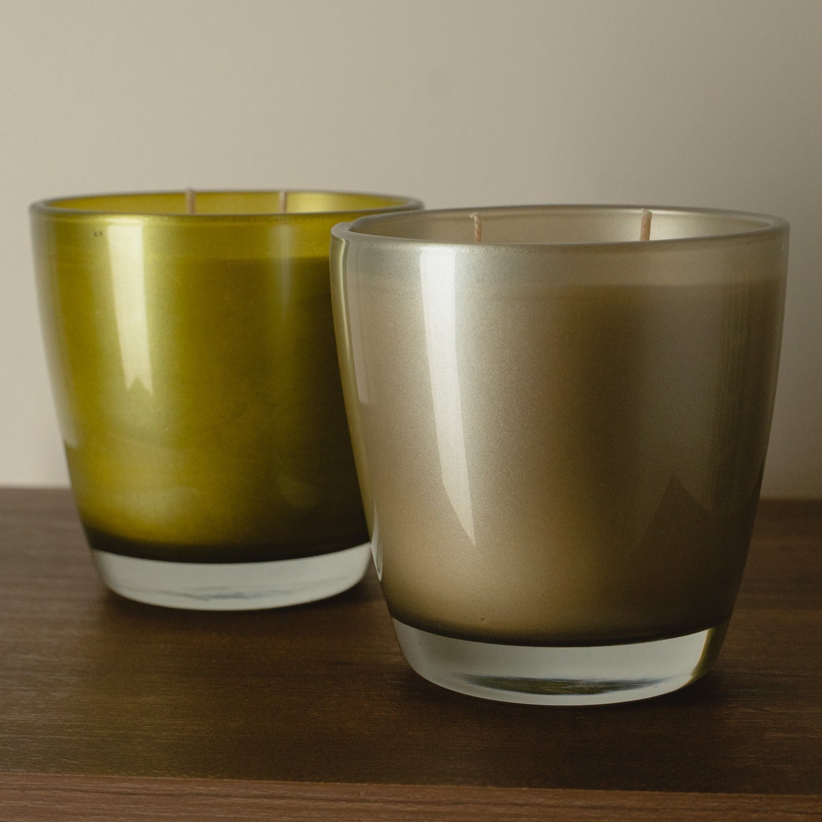 Two glass candle holders on a wooden surface against a neutral beige wall. The left holder is olive green, and the right holder is a smoky taupe-brown gradient.