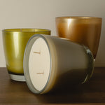 Three glass jar candles on a wooden surface against a plain beige wall. One candle is lying on its side in the foreground, showing a white wax surface and two wicks, while the other two stand upright behind it in translucent olive-green and warm amber-brown glass.