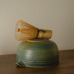 Green ceramic whisk holder with a bamboo whisk on a wooden surface