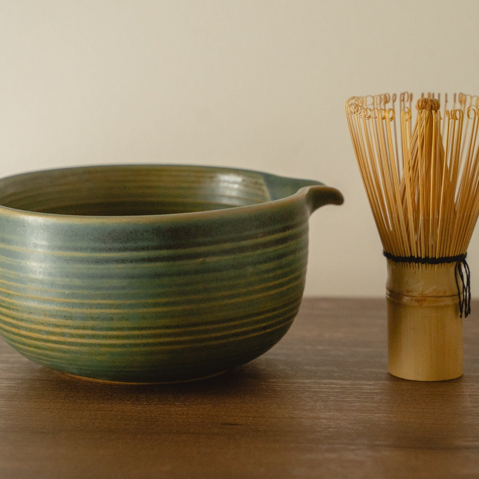 Green ceramic bowl and bamboo whisk on a wooden surface with a beige background