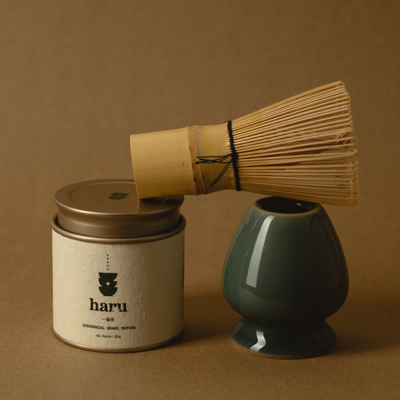A tea preparation set on a warm brown background, featuring a white jar labeled 'haru' with a gold lid, a bamboo whisk resting across the top, and a glossy dark green ceramic vase.