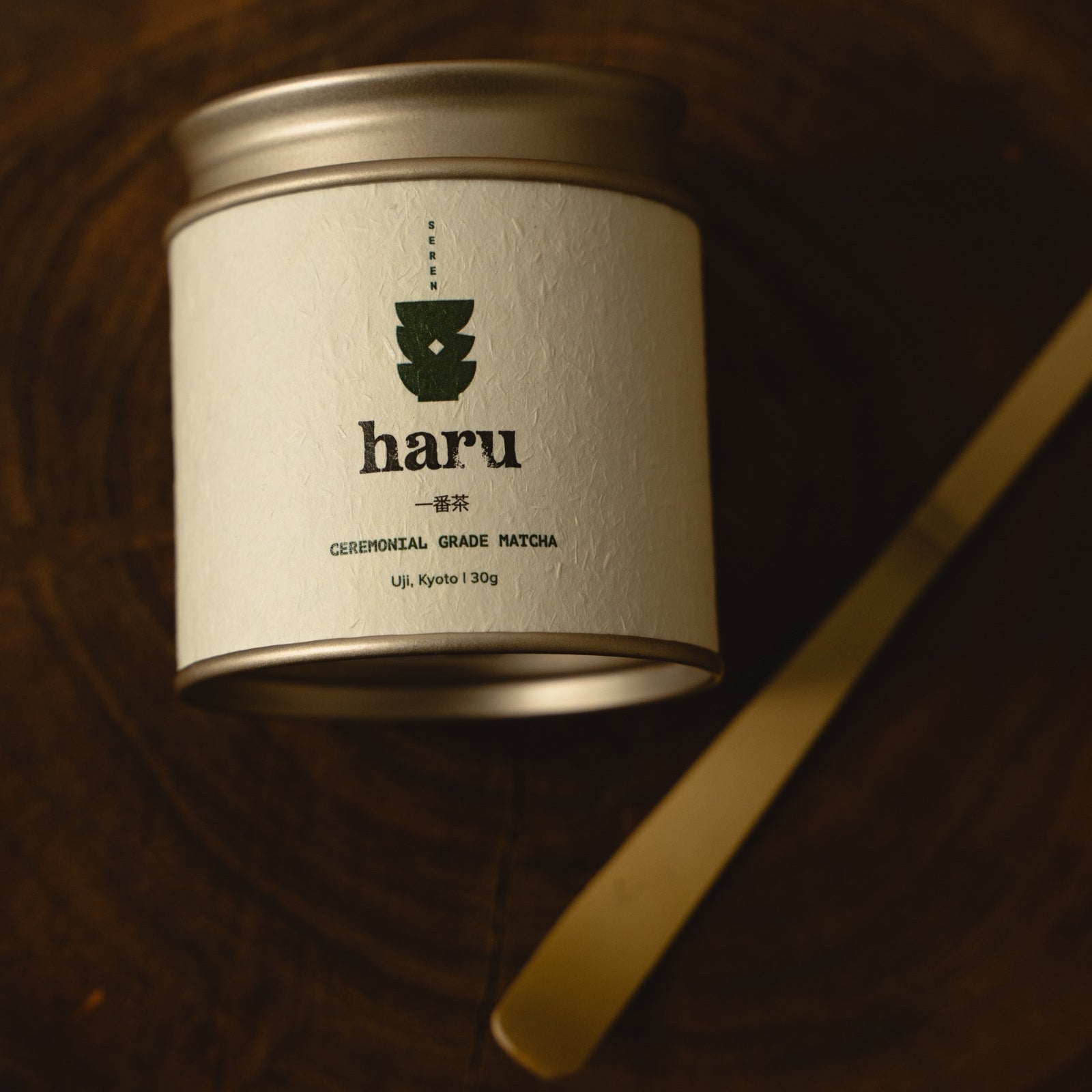 A silver metal tin of haru ceremonial grade matoya coffee, resting on a dark wooden surface next to a gold-colored spoon.
