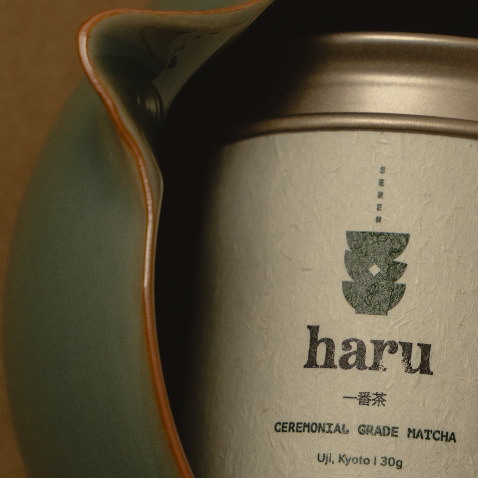 A close-up of a silver canister of haru Ceremonial Grade Matcha tea nestled inside a dark green ceramic bowl with a light orange rim, on a warm brown background.