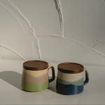 Two ceramic mugs with brown lids on a light surface against a textured white wall.