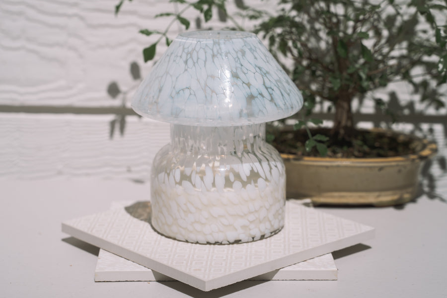Mushroom candle lamp with white spots on clear glass. Filled with 100% soy wax on tile with bonsai in the background.