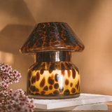 Mushroom candle lamp with light and dark brown spots on tan coloured glass. Leopard candle lamp is filled with 100% soy wax sitting on white tiles next to dried flowers.