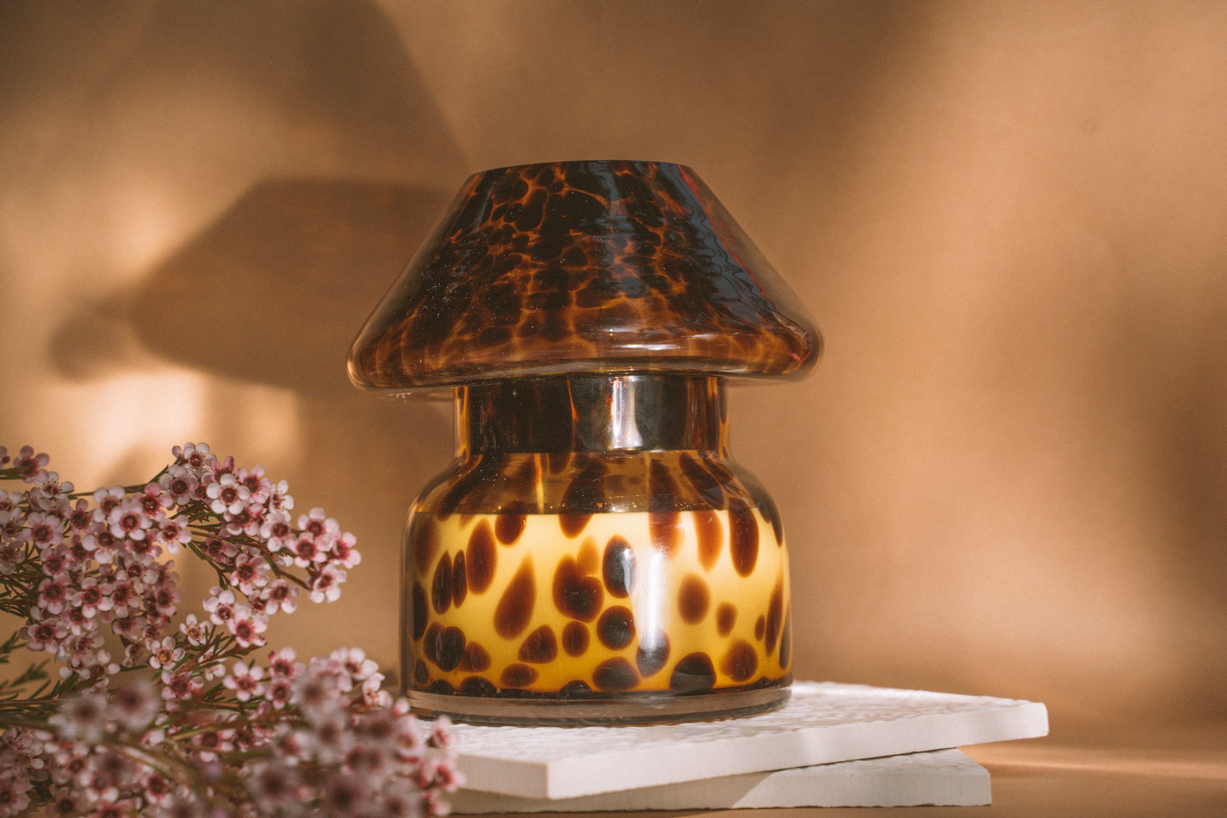 Mushroom candle lamp with light and dark brown spots on tan coloured glass. Leopard candle lamp is filled with 100% soy wax sitting on white tiles next to dried flowers.