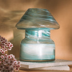 Mushroom candle lamp with aqua blue and gold glitter swirls on clear glass. Mushroom lamp is filled with 100% soy wax placed on white tiles and tan background.